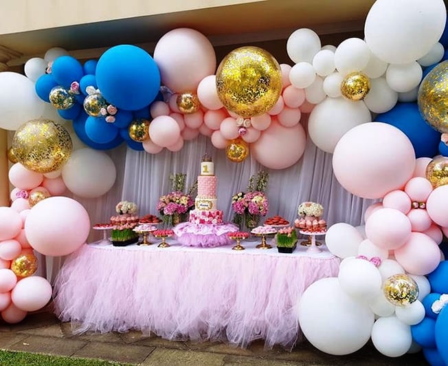 Masă de candy bar cu ghirlandă mare de baloane albastre, albe, roz și aurii, fundal roz.